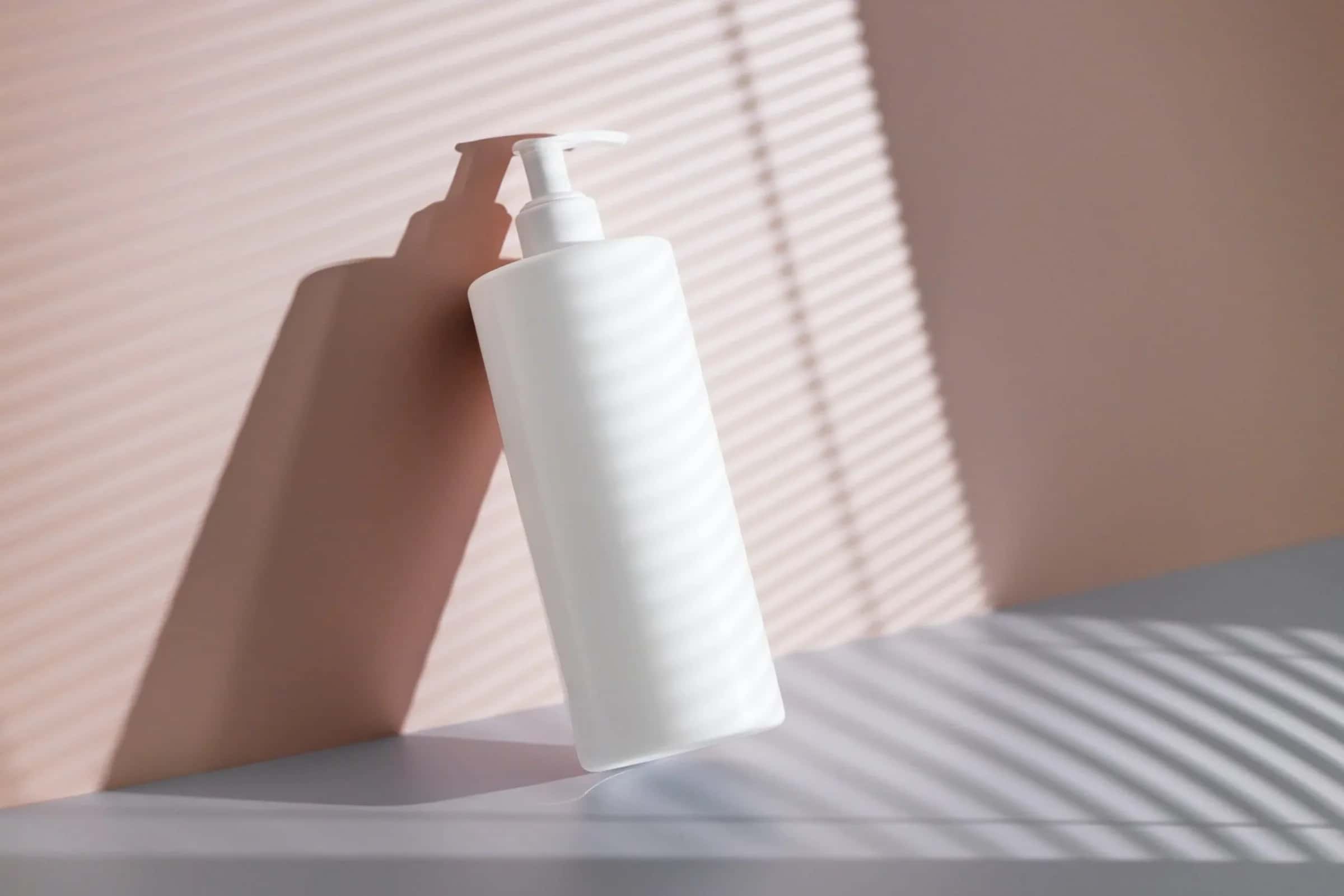 A white pump bottle leaning against a pink wall striped with window-blind shadows