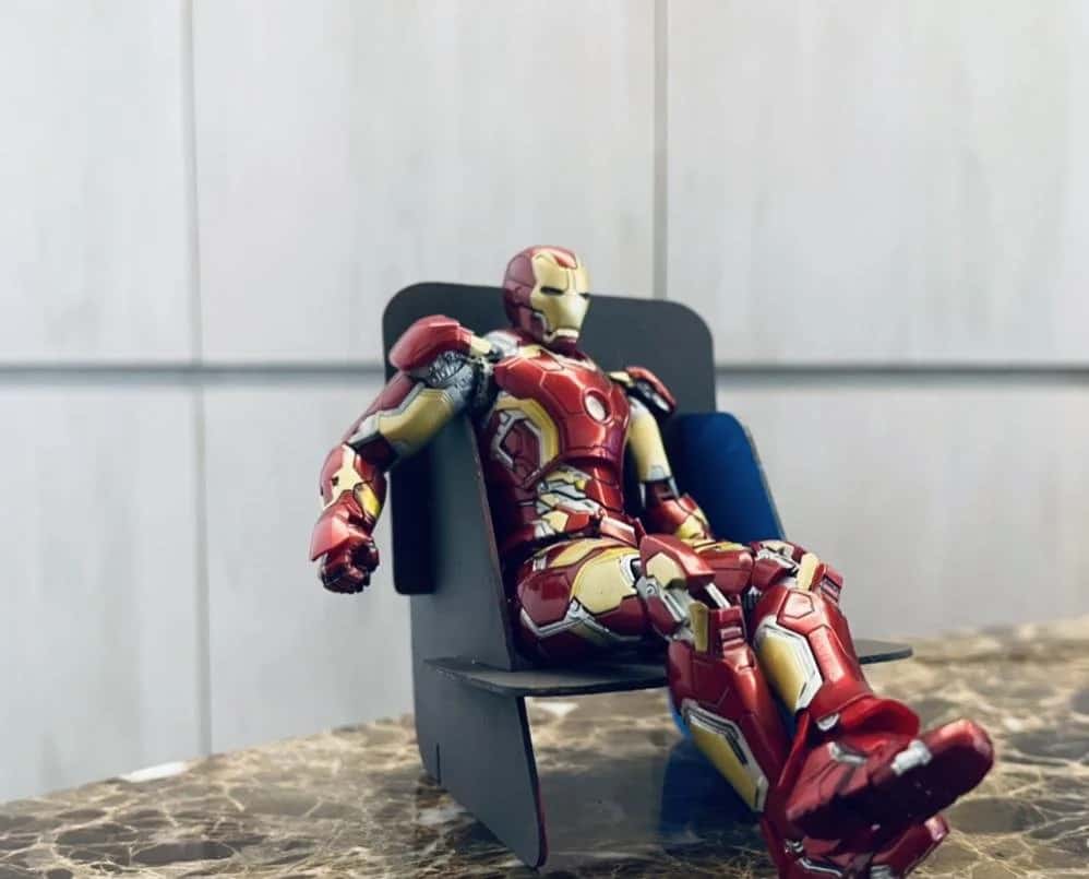 Scale model of the flat-pack chair in black board with a blue backrest, on a marble counter, with an Iron Man action figure lounging in it.