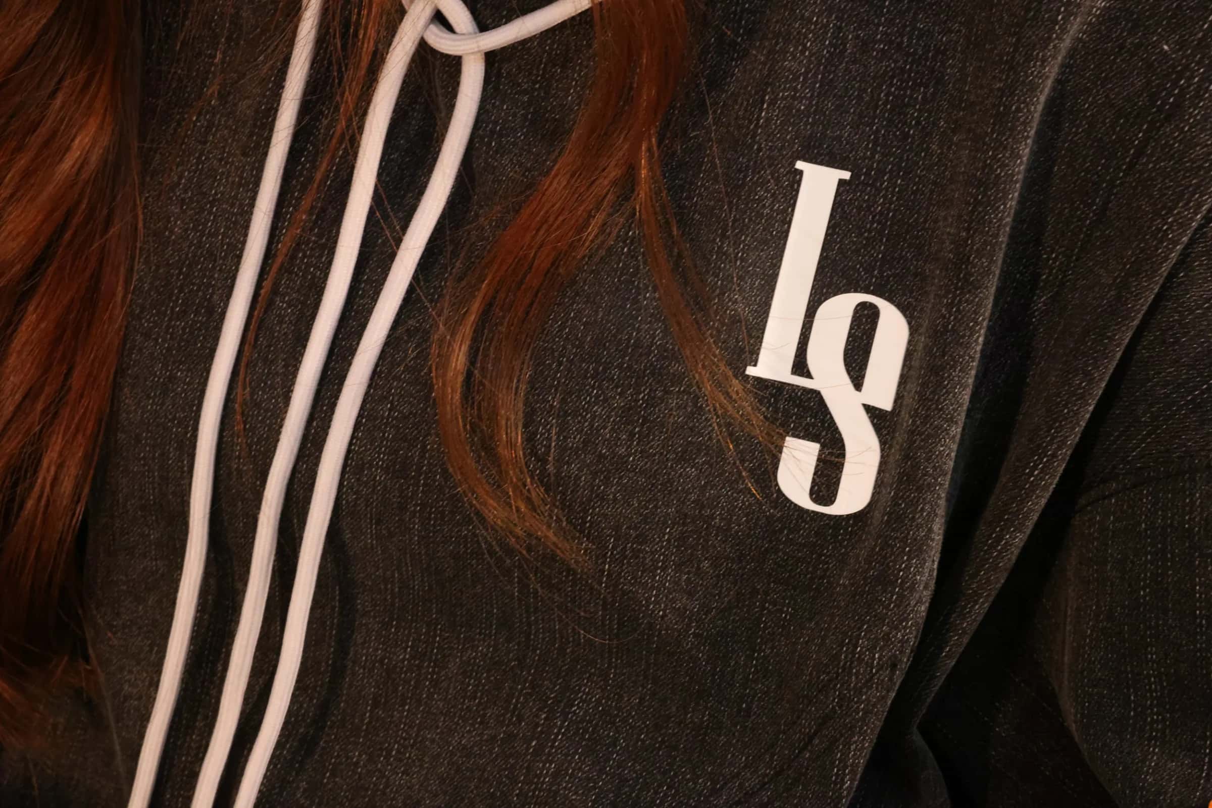 Close-up of the washed denim hoodie: a white LS monogram on charcoal denim, white drawstrings and a fall of auburn hair