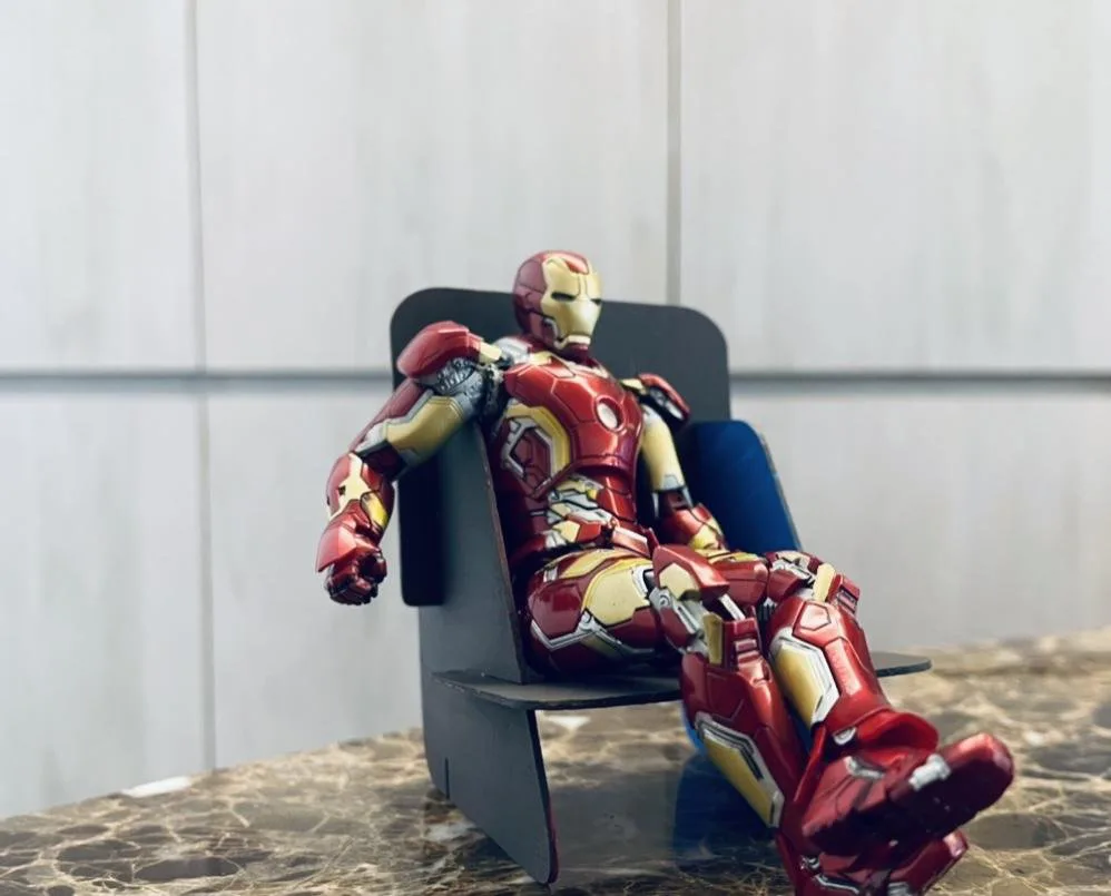 Scale model of the flat-pack chair in black board with a blue backrest, on a marble counter, with an Iron Man action figure lounging in it.