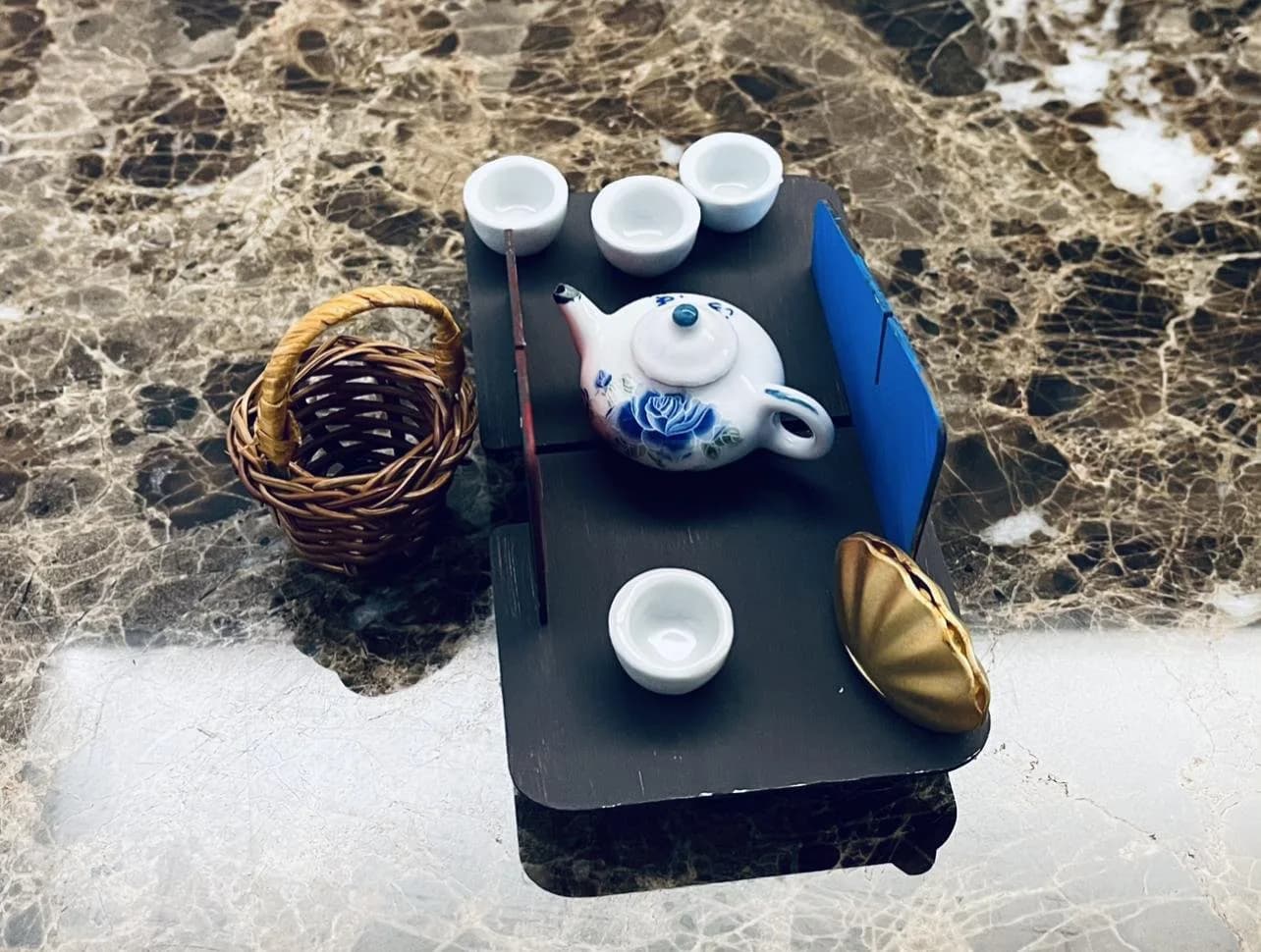 Top-down photo of the scale model in table mode on marble, set with a blue-and-white miniature tea set, a wicker basket and a shell-shaped dish.