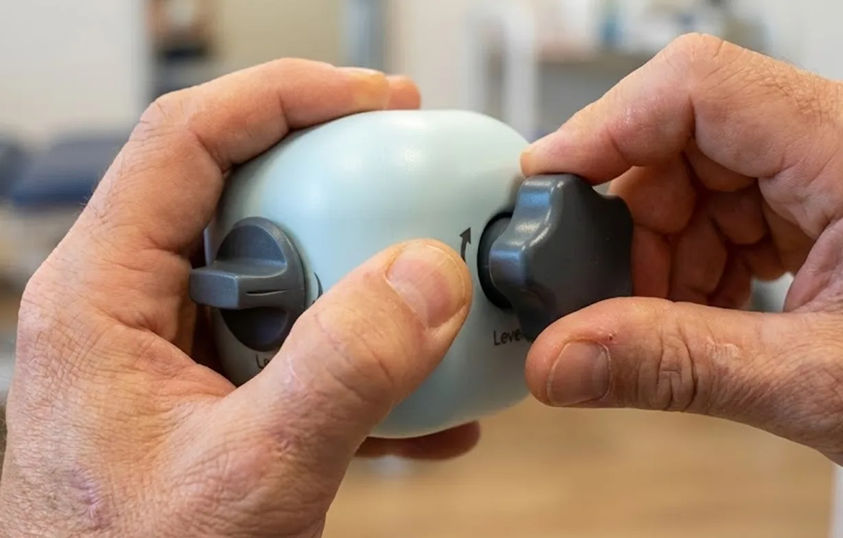 Version 1 held in two hands: one hand stabilises the cube while the other pinches and turns a lobed knob; a pointer knob is fitted on the other face and a level marking is visible.