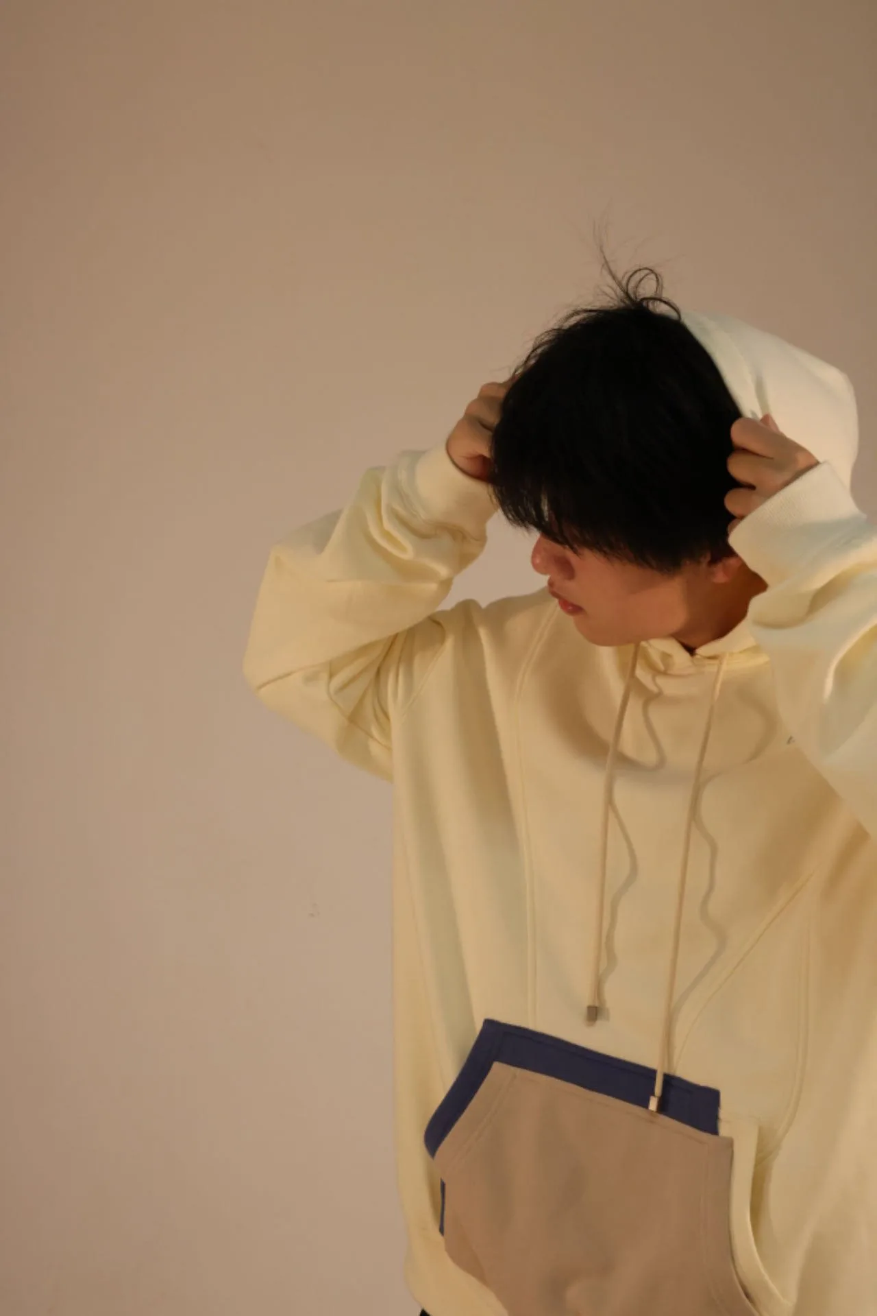Model with hands in hair wearing the cream hoodie with its blue-and-taupe front pocket, against a warm beige backdrop