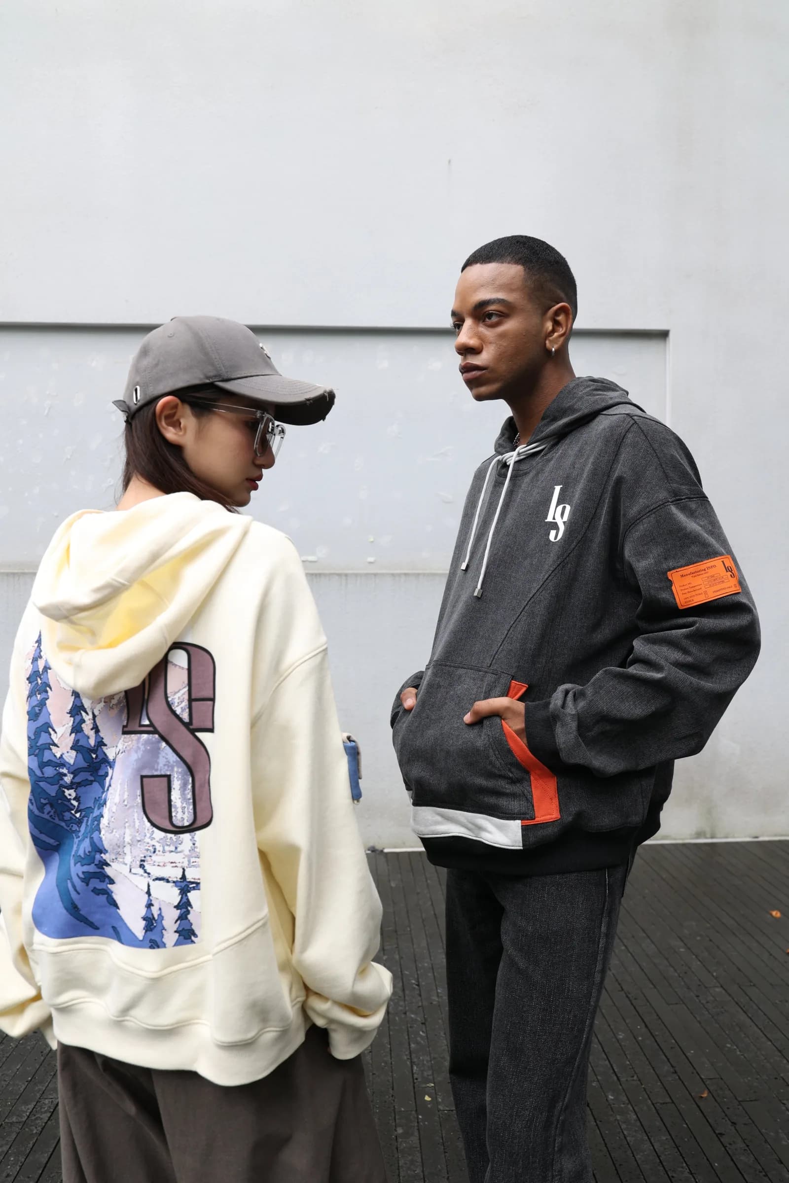 Two models against a grey wall: the cream hoodie with its snow-mountain LS back print and the washed denim hoodie with its orange label
