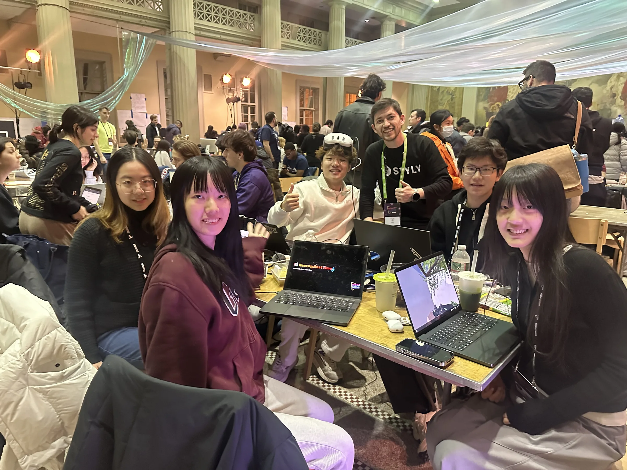 Team photo at MIT Reality Hack: teammates smiling around a table of laptops in a busy, decorated hall, a Quest headset pushed up on one teammate’s head.