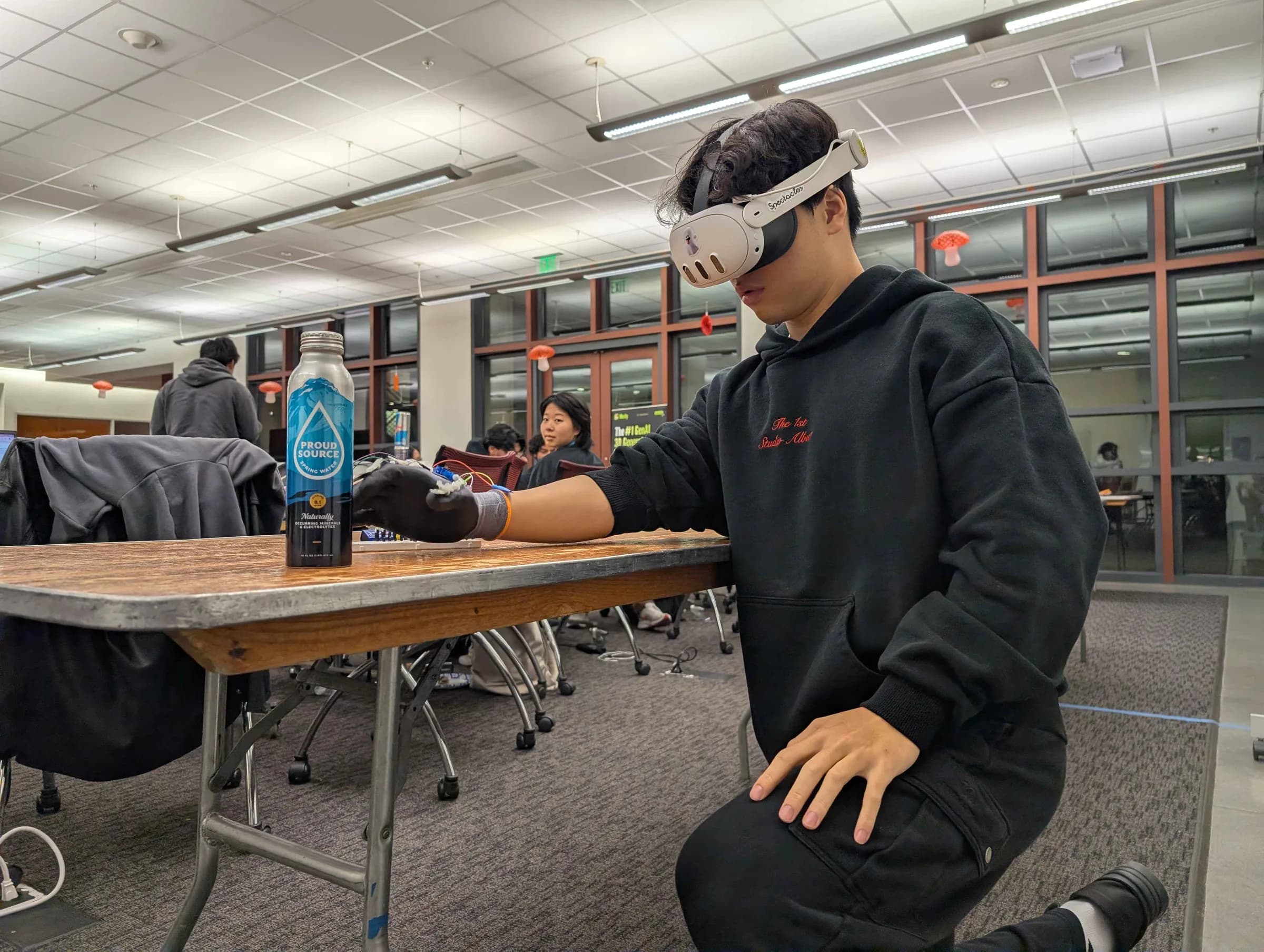 A teammate in a Meta Quest headset kneels at a table in a large study hall, his hand in the sensor glove, reaching for a water bottle.
