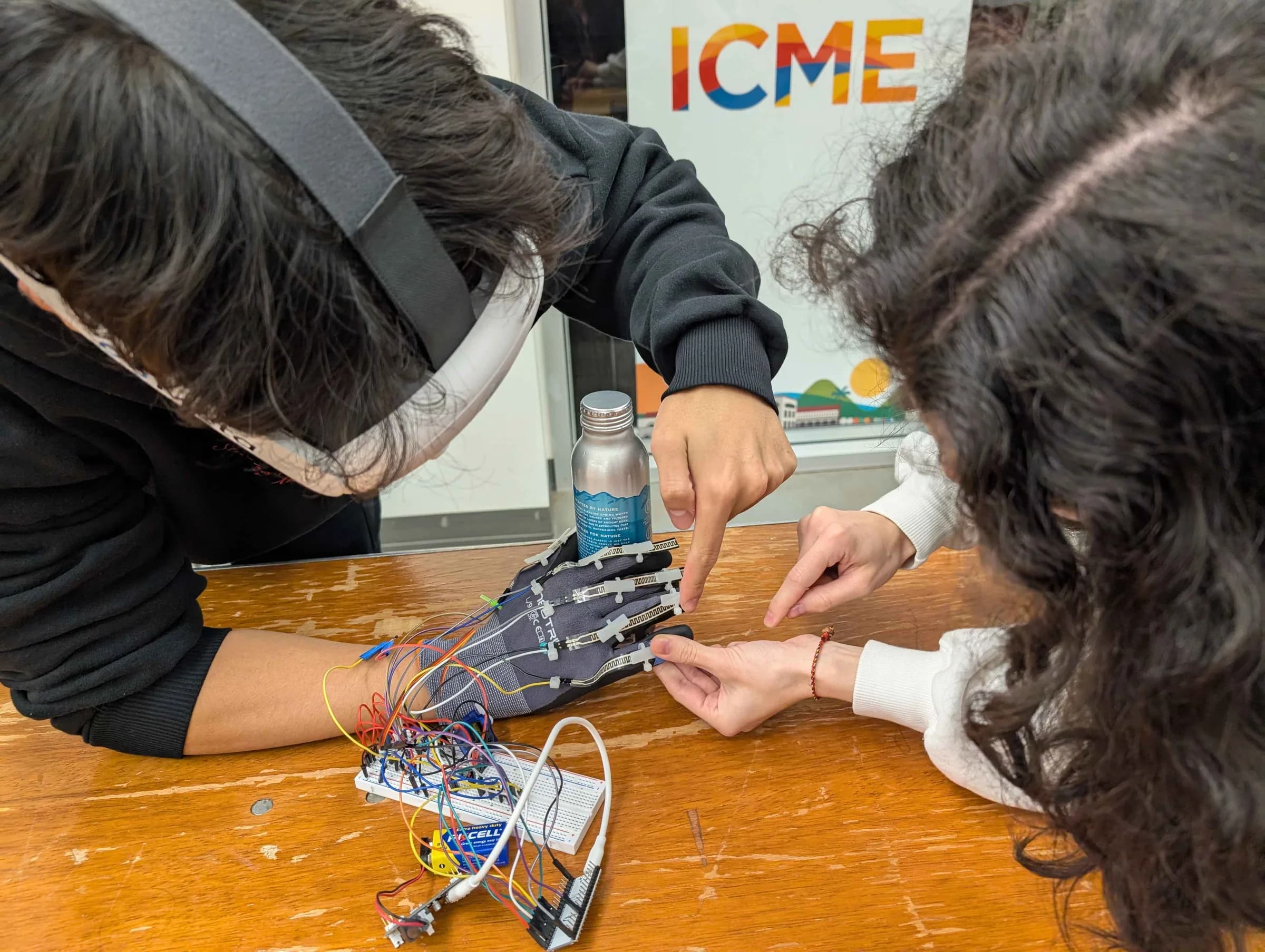 Two team members lean over a wooden table, adjusting the flex sensors on a gloved hand wired to a breadboard.