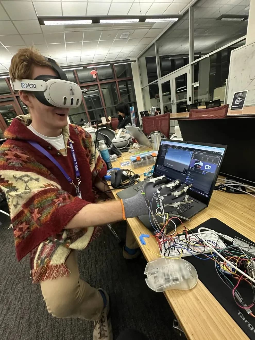 A participant wearing a Meta Quest 3 and the sensor glove kneels at a desk, smiling, beside a laptop running the Unity scene and the wired breadboard.