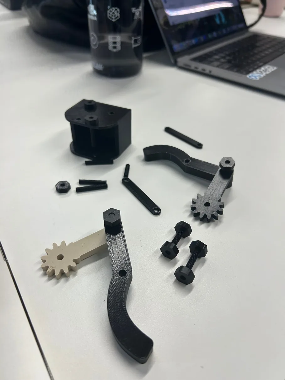 Early black and tan test prints on a desk: gears, a curved finger, a servo housing and short links laid out before assembly