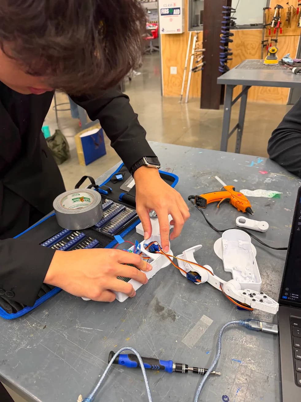 Hands holding white printed arm parts together on a blue silicone mat, with a hot-glue gun and tape nearby