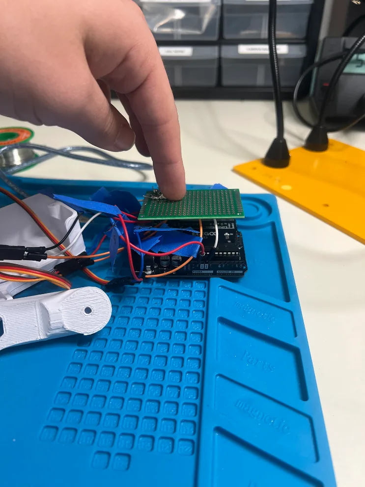 A finger pressing a green soldered protoboard onto an Arduino on a blue silicone work mat, servo wires attached