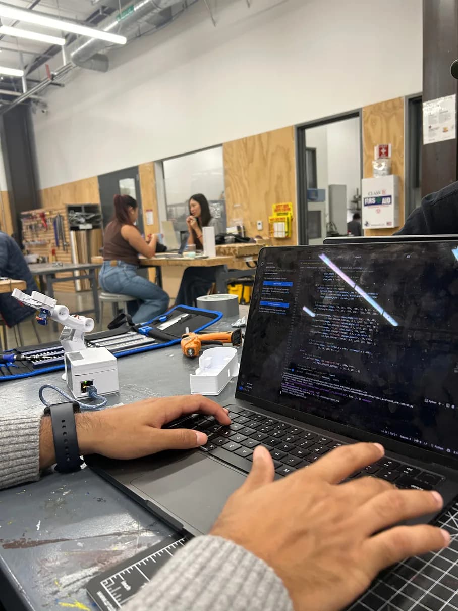 Over-the-shoulder view of hands typing code on a laptop in a busy makerspace, the white arm on the bench to the left