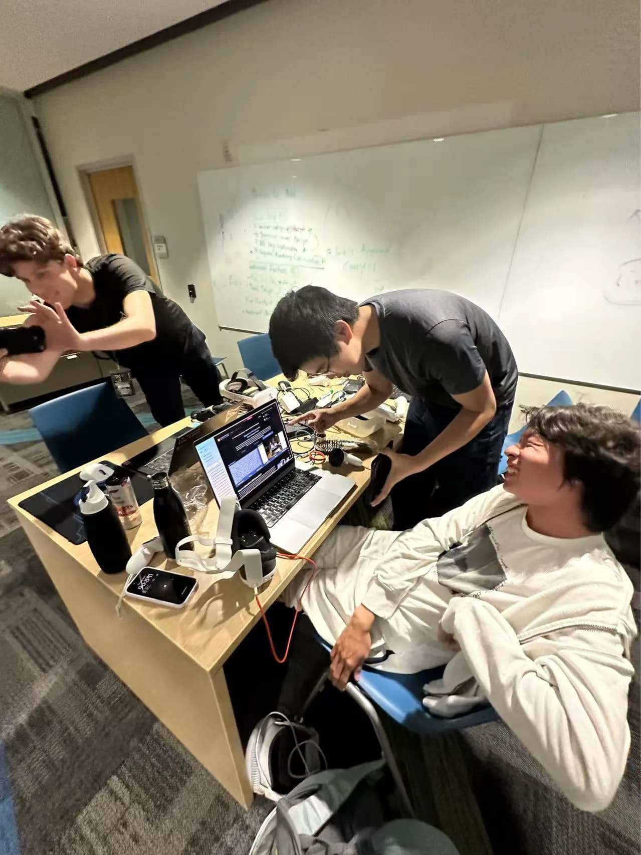 Hackathon photo: three teammates crowd around a laptop, one leaning back laughing at a breakthrough.