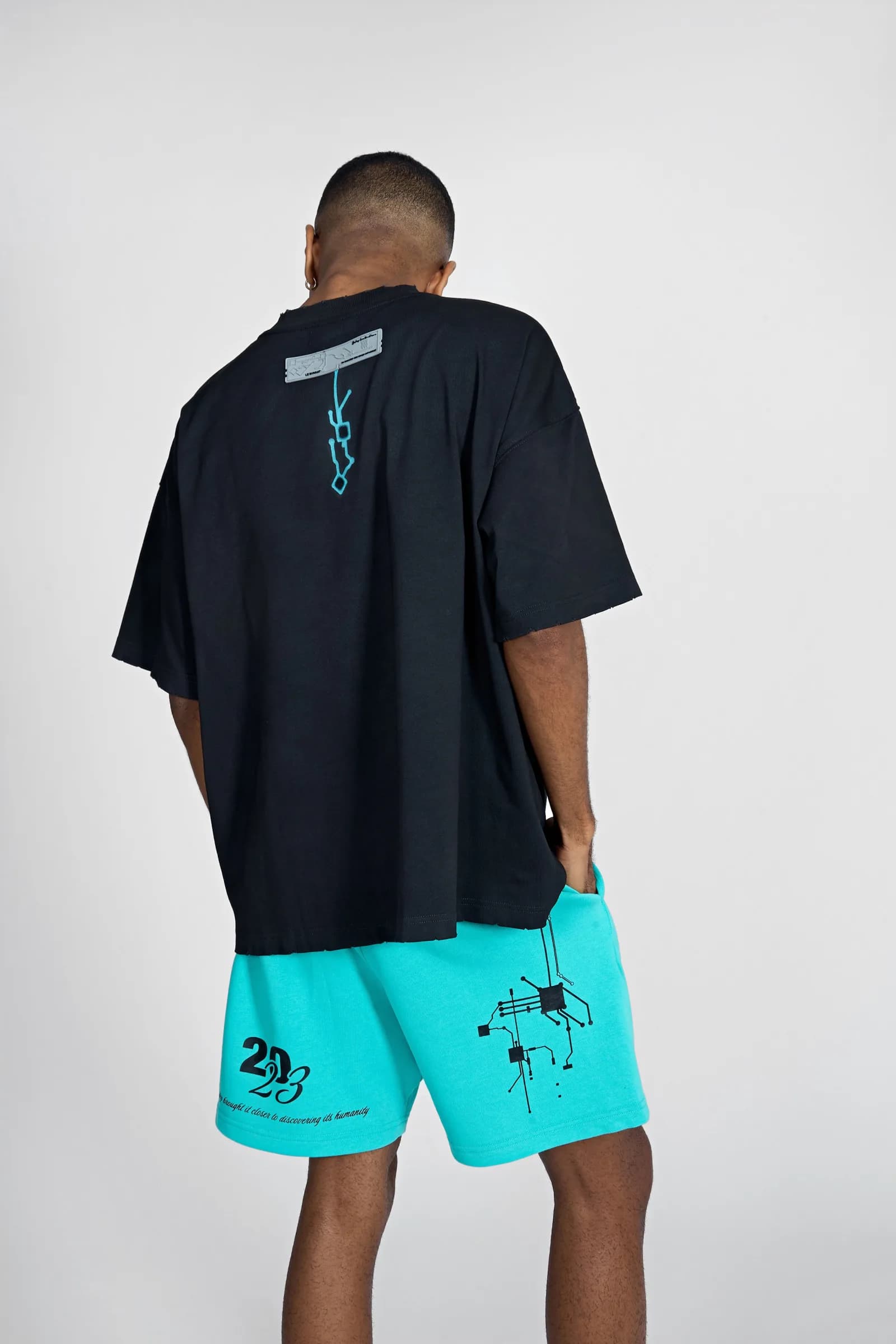Model from behind: the Birth tee back print with cyan shorts showing the 2023 script and motherboard graphic