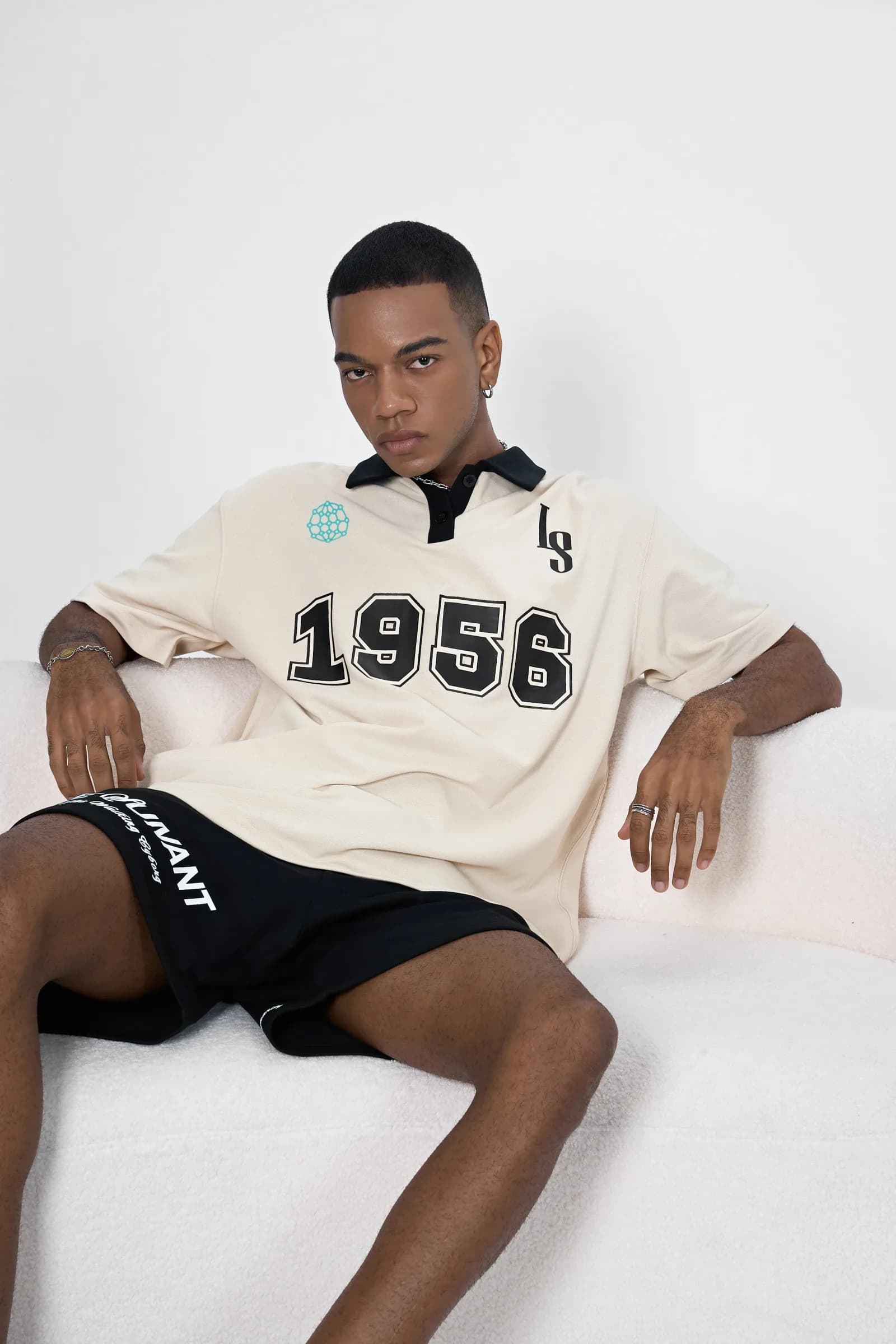 Model seated on a white block in the Butterfly Effect polo with 1956 numerals and black Le Suivant shorts