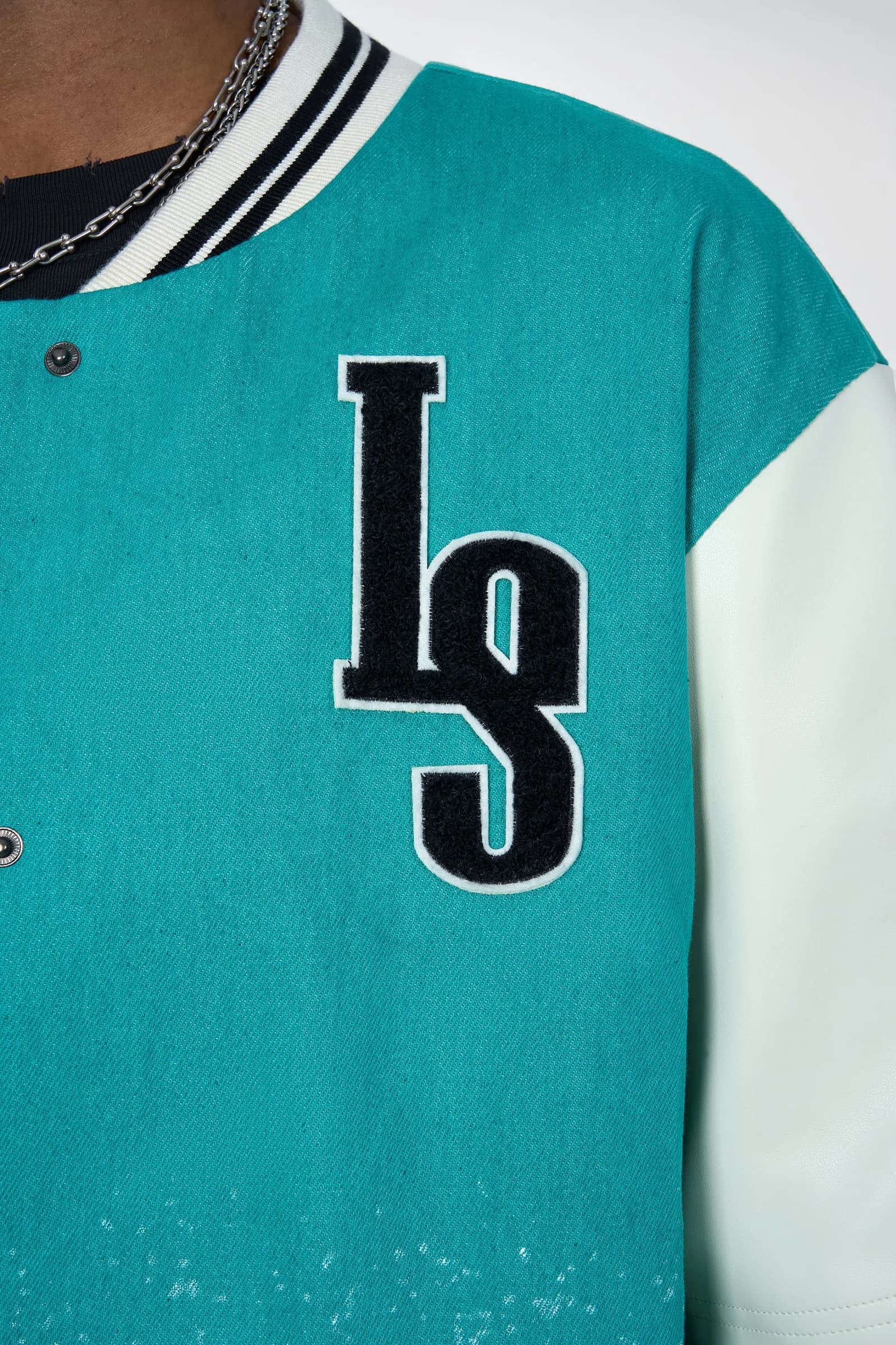Close-up of the embroidered black LS monogram on the cyan denim chest, with a cream sleeve and striped rib collar