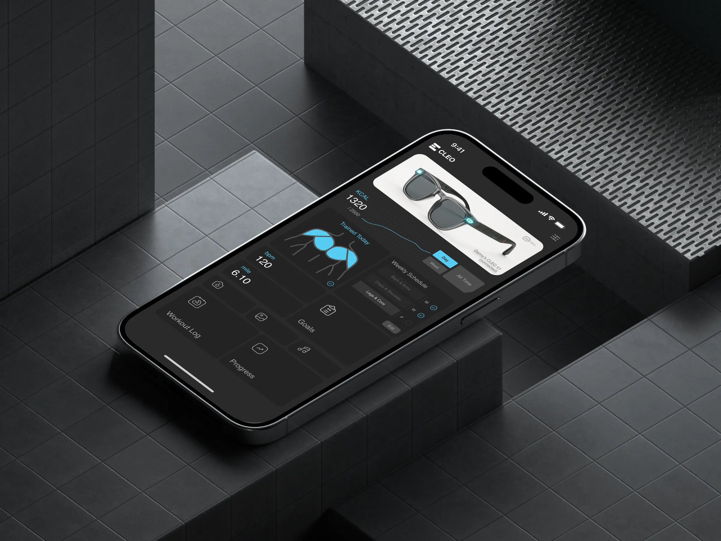 Companion app mockup: a phone on dark tiled blocks running the CLEO app — a “Danny’s CLEO V1 (connected)” glasses card, a KCAL 1320/2500 chart, a “Trained Today” muscle map, bpm and mile stats, a weekly schedule and Workout Log, Goals and Progress tiles.