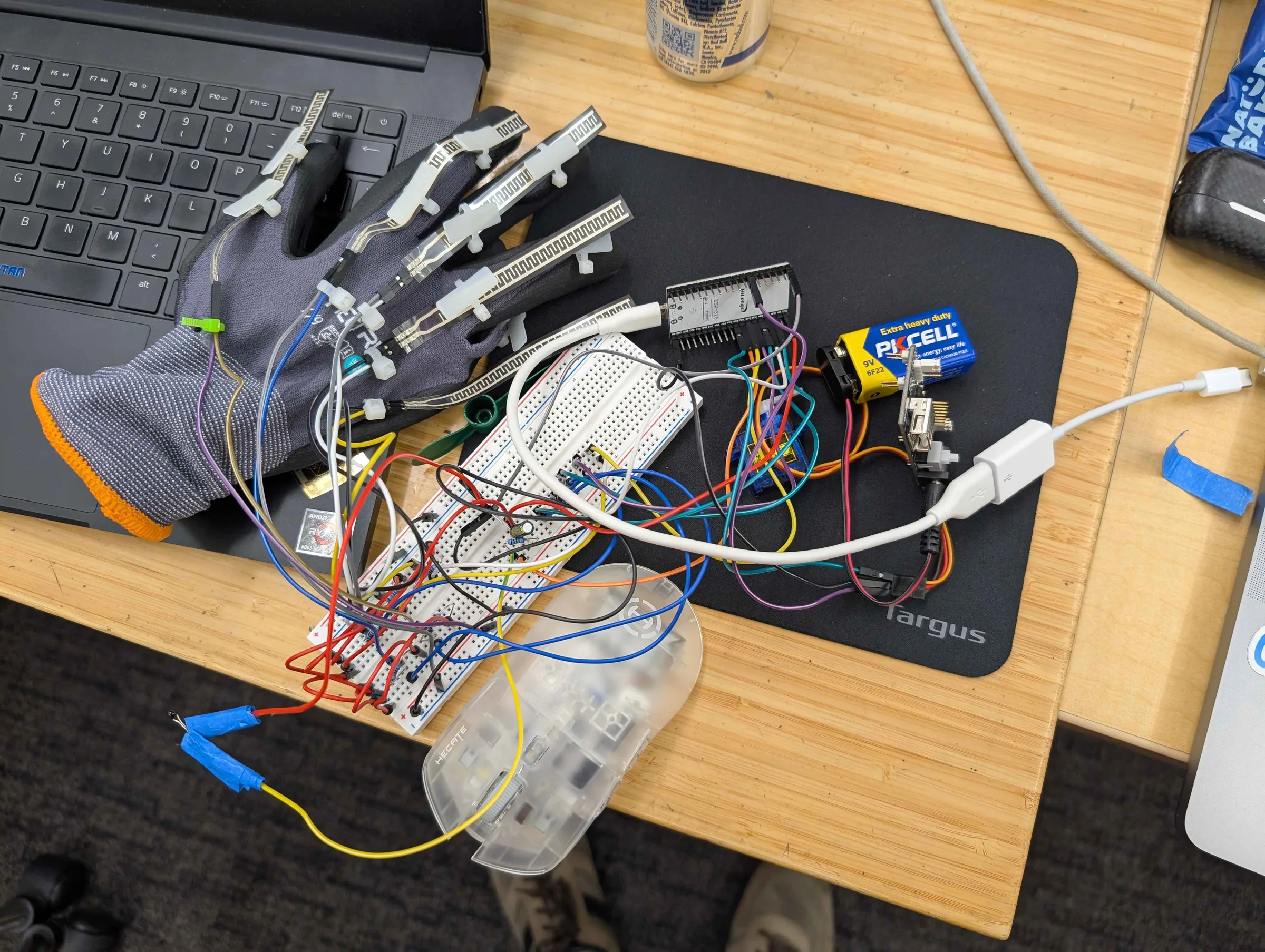 The DIY FlexGlove on a desk: a grey work glove with a flex sensor zip-tied along each finger, jumper wires running to a breadboard, an ESP32, a servo and a 9V battery, beside a laptop.