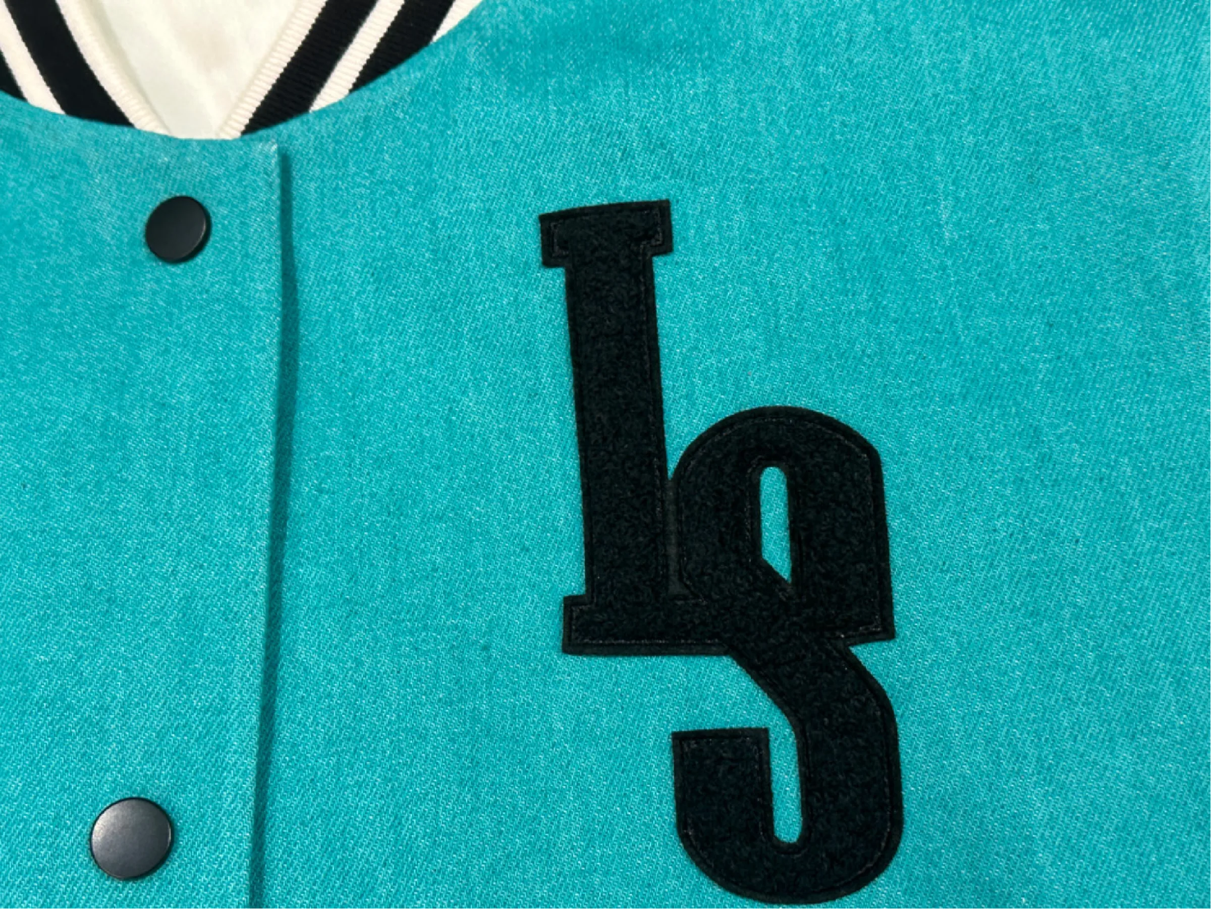 Macro photo of the black embroidered LS monogram and a snap button on the cyan denim of the produced bomber
